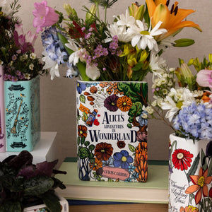 Alice in Wonderland ceramic book vase used as flower holder on bookshelf with other literary vases.