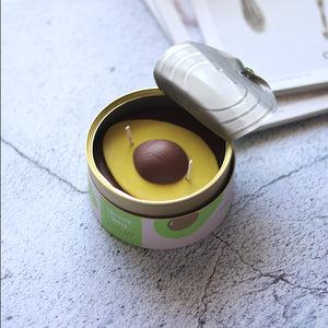 Side angle of the fresh herb-scented avocado candle with open tin lid, styled on a textured surface next to a magazine.
