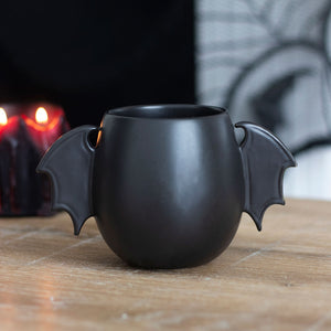 Black bat wing Halloween mug on wooden table with candlelight, featuring curved ceramic bat wings and matte black finish – gothic home décor style.