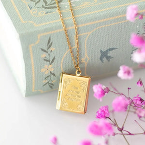 Gold book-shaped locket necklace engraved with “You are my favourite chapter”, shown styled with a pastel book and pink flowers.