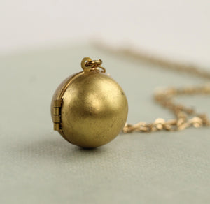A close-up of a vintage brass sphere locket necklace resting on a soft green background, showcasing its smooth, round shape and antique finish.