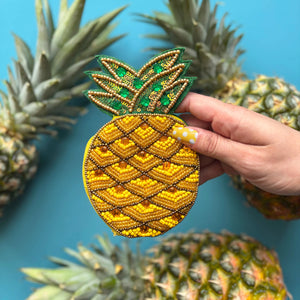 Fruity pineapple coin purse held in hand against a blue background, surrounded by fresh pineapples.