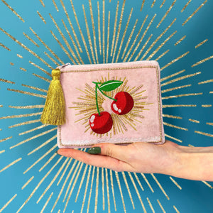 Hand holding pink velvet cherry purse with gold embroidery and tassel against a vibrant blue and gold starburst background.