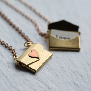 Gold envelope locket necklace with copper heart on rose gold chain, shown closed on textured background.