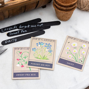 Front view of assorted vintage-style flower seed packets for daisies, forget-me-nots, and sweet peas, with chalk labels in background.