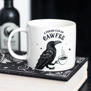 White ceramic mug with black raven holding a steaming cup and text “A Fresh Cup of Cawfee”, styled on gothic book.
