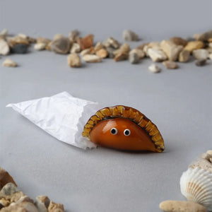 Glass Cornish pasty ornament with googly eyes peeking out of a white paper wrapper, surrounded by pebbles and seashells.