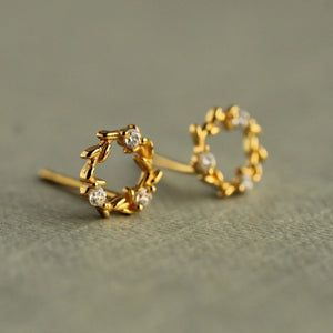 Close-up front view of gold wreath circle stud earrings with laurel leaf design and sparkling cubic zirconia stones on a soft neutral background.
