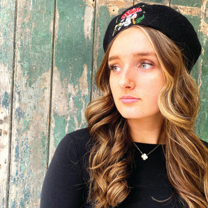 Woman wearing black wool beret with embroidered red toadstool, ferns, and stars, standing in front of distressed green wooden background – woodland fashion accessory.