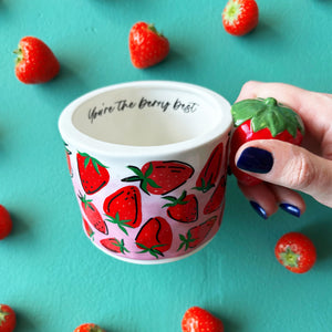 Hand holding strawberry-themed ceramic mug with 3D strawberry handle and “You’re the berry best” printed inside, surrounded by real strawberries on teal background.
