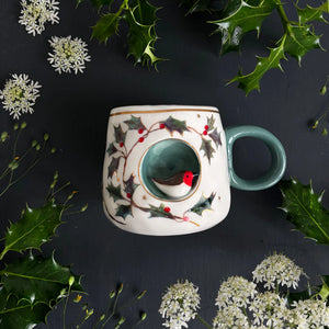 Top view of Secret Garden robin mug with 3D bird inside, surrounded by holly and white flowers on a dark background.