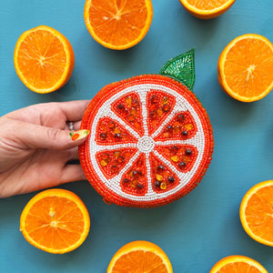 Hand holding a citrus beaded purse in front of orange halves, showing the playful fruit-inspired design and sparkle.