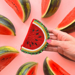 Person holding a beaded watermelon purse in front of watermelon slices, showcasing the vibrant seed pattern and curved zip design.