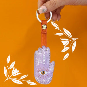 Person holding Hand of Fatima keyring with mystical eye print, styled on orange background with floral overlay.
