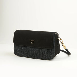 Front angle of the Caithness Clutch Bag featuring black herringbone Harris Tweed® with a soft black leather flap, gold crest emblem, and detachable braided strap.