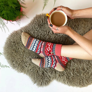Cosy Fair Isle Moomin socks worn by person holding a mug of tea on a fluffy rug, showing red and pink knit with Little My embroidery.