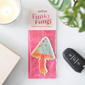 Vanilla-scented mushroom air freshener in pink packaging on white desk with candle, car key, and plant – fun and stylish car accessory.