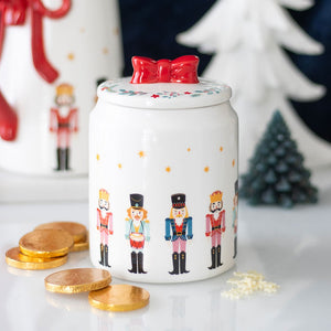 Festive ceramic nutcracker storage jar with red bow lid, displayed with chocolate coins and Christmas decor in the background.