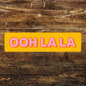 Ooh La La orange and pink metal sign with retro lettering on rustic wooden background – fun 12x3 inch aluminium wall art for indoor or outdoor use.