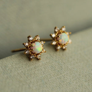 Close-up front view of gold-plated sterling silver opal flower stud earrings with white opals and sparkling cubic zirconia petals on a soft neutral surface.