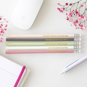 Pastel book-inspired pencils with gold-foil quotes arranged beside a notebook and flowers.