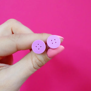 Front view of lilac button stud earrings held between fingers against a bright pink background – handmade acrylic jewellery for sewing lovers.