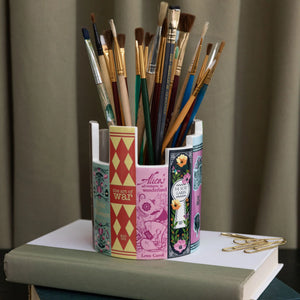 Ceramic literary book pot used as a paintbrush holder on a desk, surrounded by books, gold paperclips, and soft beige curtains.