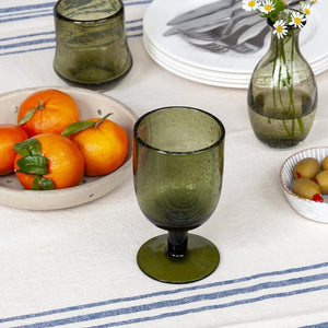 Green stemmed bubble glass on a rustic table set with fruit, olives, plates and a striped tablecloth.
