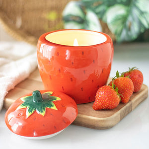 Lit strawberry-shaped candle jar with ceramic lid off, styled on a wooden board with fresh strawberries and tropical leaves in the background.