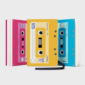 Three colourful cassette-style notebooks in pink, yellow, and blue, standing upright and designed to look like retro mixtapes.