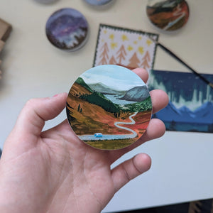 Hand holding a round vinyl sticker featuring a painted Highland landscape with a winding road and a small blue camper van, surrounded by mountains and moorland.