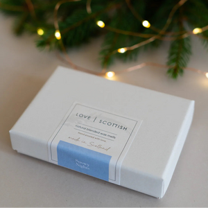 Snowy Nights wax melts gift box by Love Scottish with soft fairy lights and evergreen branches in the background.