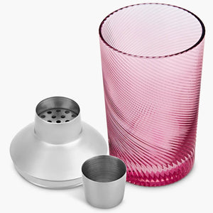 Disassembled pink glass cocktail shaker showing stainless steel lid, strainer, and cap beside the twisted textured glass base.