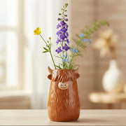 Brown ceramic Highland Cow vase with horns holding purple delphiniums and yellow flowers on wooden surface
