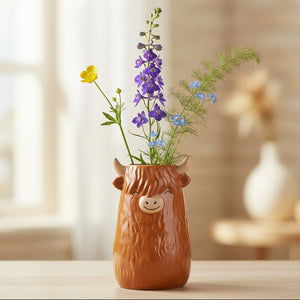 Brown ceramic Highland Cow vase with horns holding purple delphiniums and yellow flowers on wooden surface