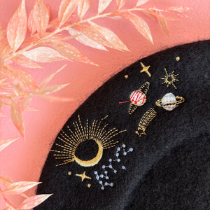 Close-up detail of gold embroidered moon and planets on black wool beret with pink background.