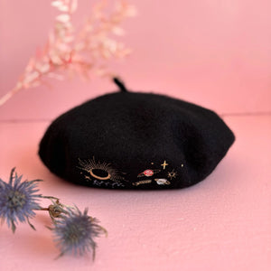 Black beret with embroidered galaxy design displayed on pink background.