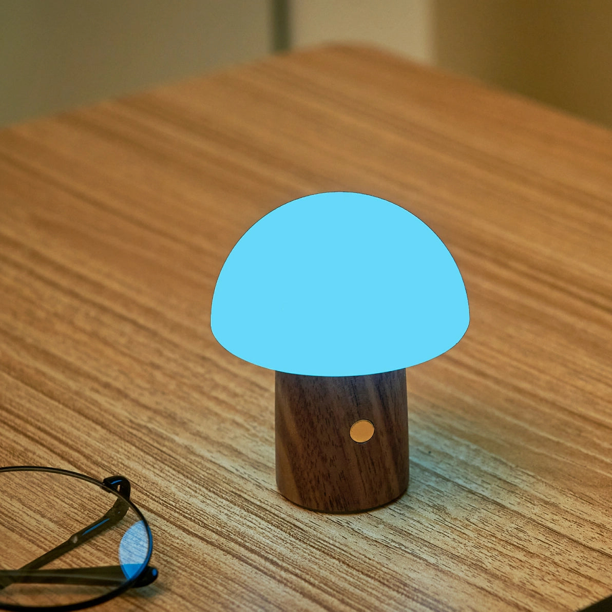 Blue glowing Alice Mushroom Lamp Super Mini on a wooden table beside glasses.