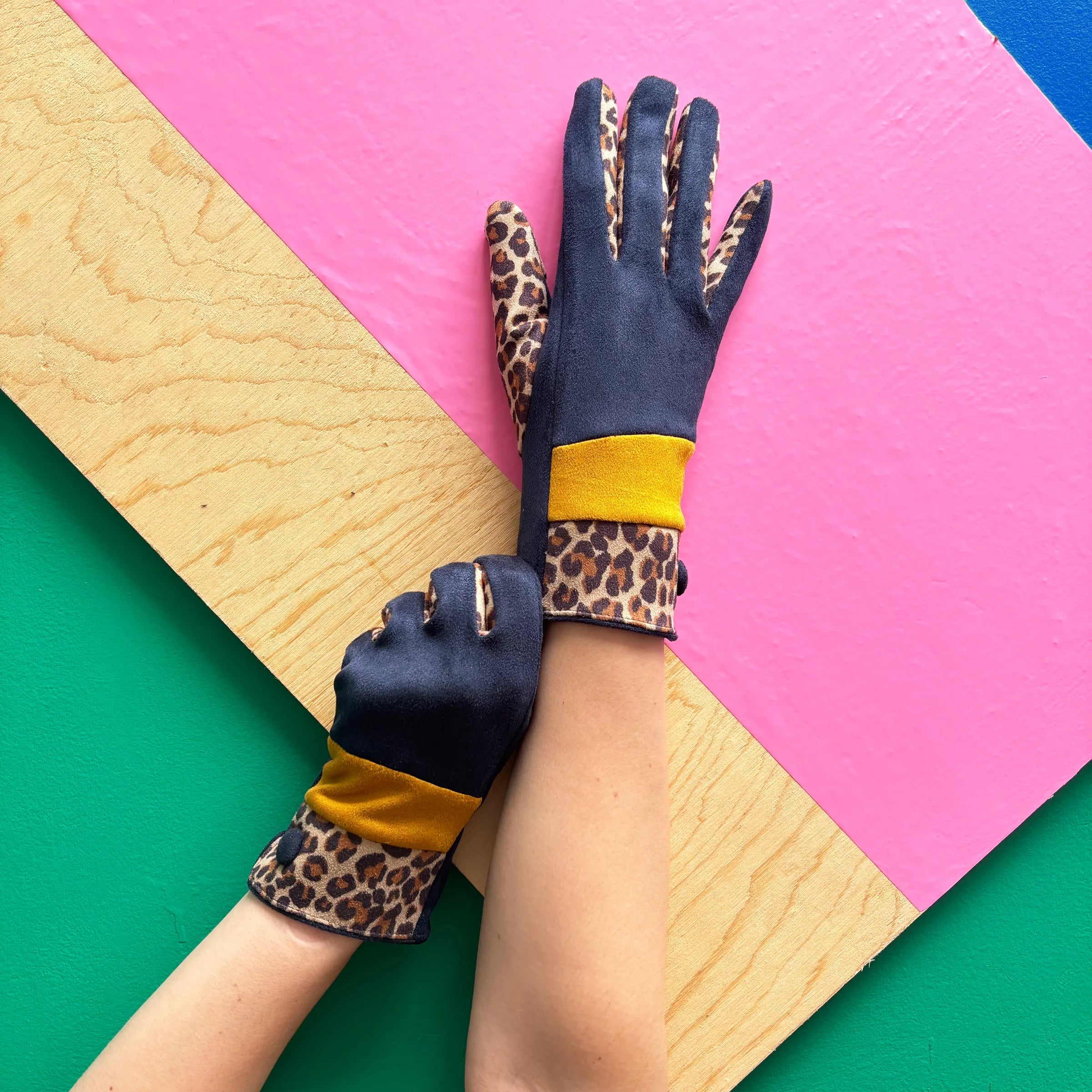 Stylish navy suedette gloves with leopard print trim and ochre band, model wearing gloves on pink and green background.