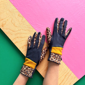 Pair of navy suedette gloves with ochre yellow accent and leopard print cuffs, displayed on model’s arms.