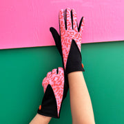 Hands wearing animal print pink and black gloves with bold leopard pattern and orange button detail, styled against a pink and green background.