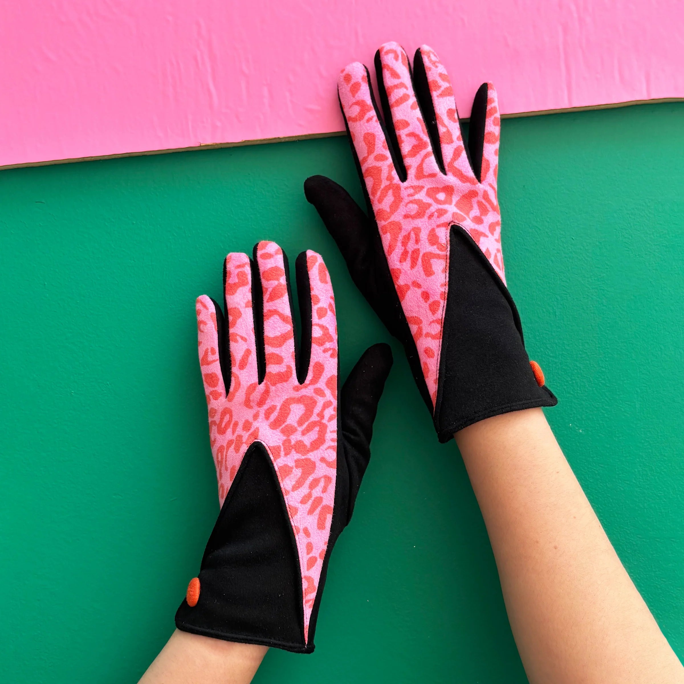 Pair of pink leopard print gloves with black contrast panels and orange button, displayed on hands against colourful background.