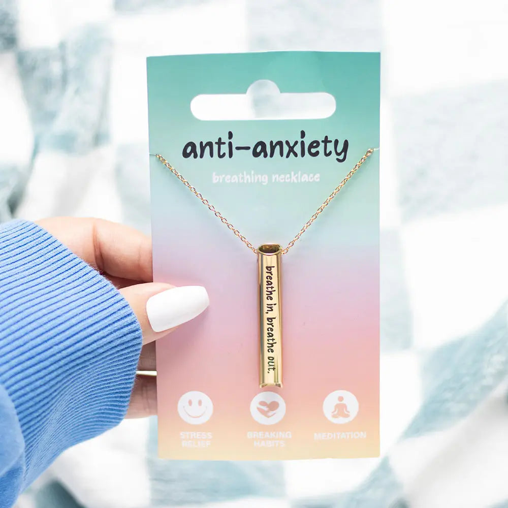 Person holding gold anti-anxiety breathing necklace on pastel packaging that reads “breathe in, breathe out” for stress relief and mindfulness.