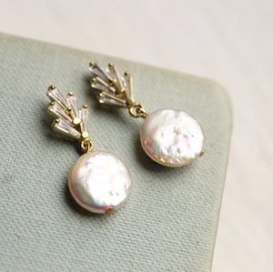 A pair of Art Deco crystal pearl earrings resting on a textured light green fabric. The earrings feature a geometric crystal design with a round, iridescent freshwater pearl.