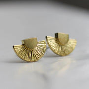 A pair of gold Art Deco stud earrings with a geometric fan design, displayed on a neutral background. The textured brass surface catches the light.