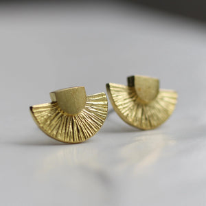 A pair of gold Art Deco stud earrings with a geometric fan design, displayed on a neutral background. The textured brass surface catches the light.