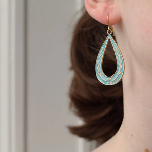Brass teardrop hoop earring with hand-enamelled floral sprig design in cornflower blue, shown hanging just below the earlobe on model.