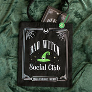 Black tote bag with “Bad Witch Social Club” design and matching pin badge on green fabric.