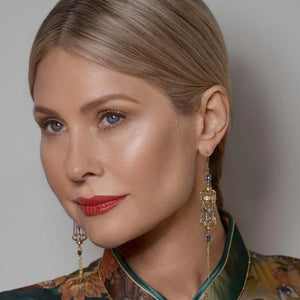 Close-up of model wearing gold bird cage earrings with blue crystals and miniature blue bird detail, styled with sleek hair and bold red lipstick.