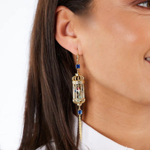 Side view of gold-plated bird cage earring with hanging chain detail, blue crystal embellishments, and enamel bird, worn by brunette model.